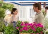 Emily em Paris chega à última temporada; Netflix divulga primeiras imagens e novidades "A woman and man talk and smile at an outdoor flower market, standing among vibrant purple flowers and green plants under a striped canopy, with a white building in the background."